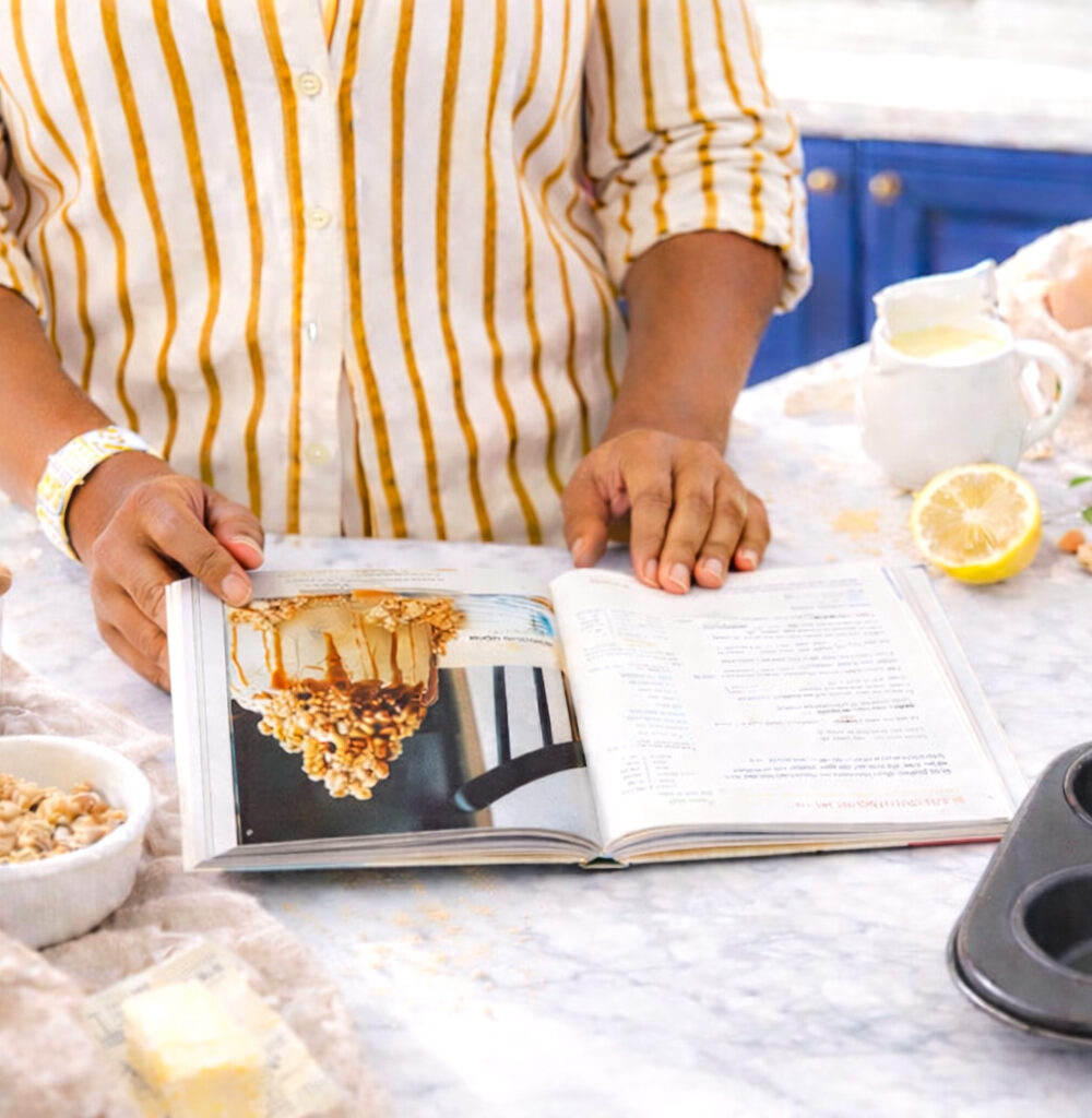 Woman preparing to bake in a bright kitchen, standing at a white marble counter with ingredients laid out, a muffin pan nearby, and an open cookbook, with deep blue cabinets in the background.